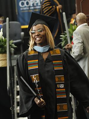 一名棕色皮肤的女子戴着眼镜，身穿黑色毕业袍，戴着帽子，穿着肯特布，右手拿着学位证书，悄悄地从毕业台上走了出来