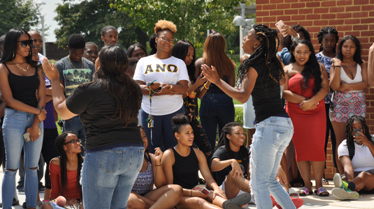 一群不同的学生站在外面观看穿着黑色t恤和牛仔裤的学生表演。