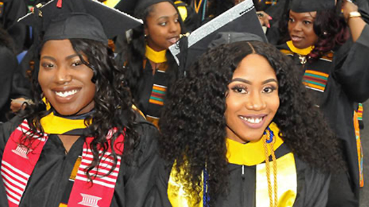 2个微笑的学生戴着黑色的毕业帽和毕业服，穿着彩色的披肩。