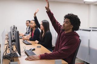 学生们坐在计算机实验室的工作站前。两个学生举手提问。
