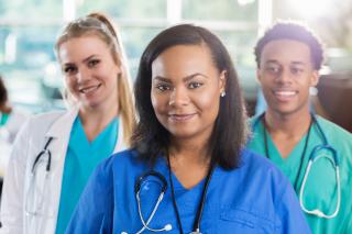 年轻的非洲裔美国女护士站在白人医生和非洲裔美国男护士面前。他们穿着手术服和白大褂，看着摄像机。